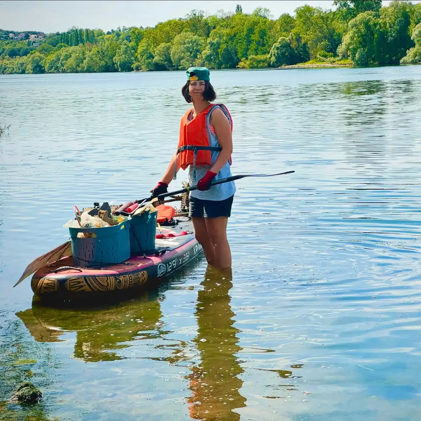 Bénévole debout dans la Seine à côté de son paddle chargé de deux bacs de déchets
